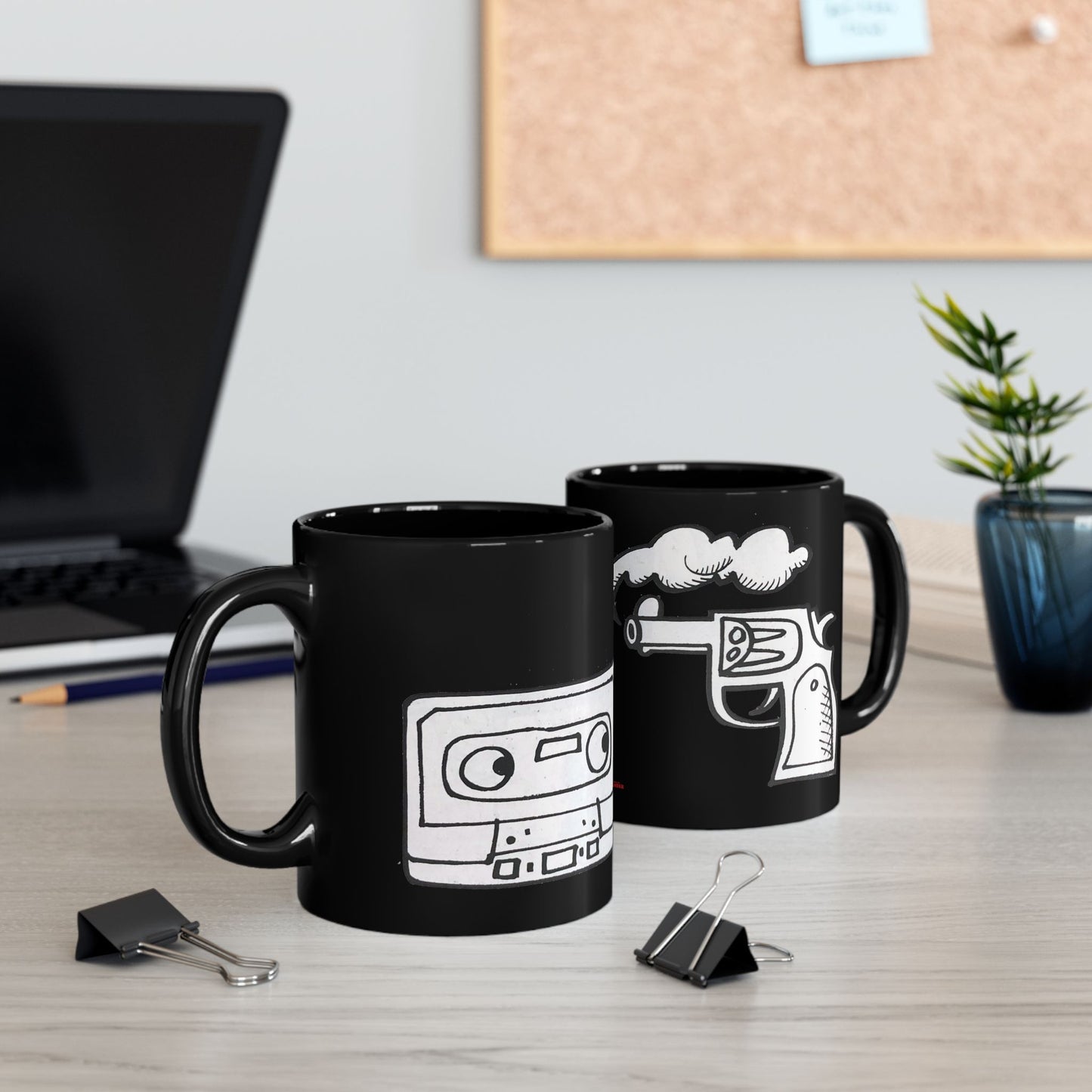 Two black ceramic mugs on a desk featuring a hand-drawn cassette and a smoking revolver illustration.