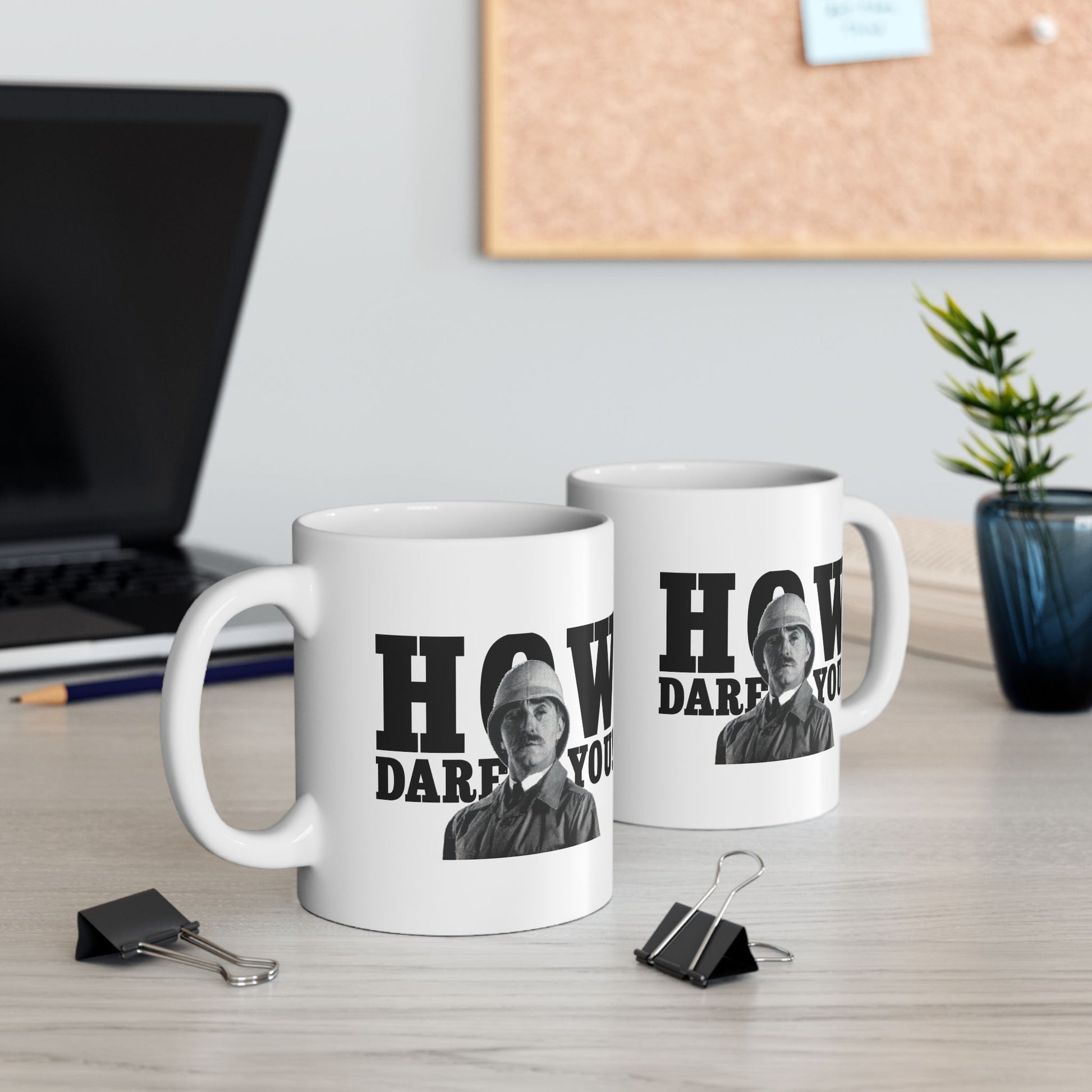Two white ceramic mugs featuring a stern British soldier with the phrase "How Dare You" in bold text, displayed on an office desk.