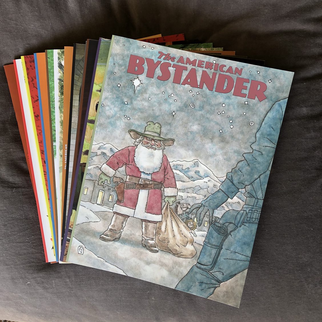 A stack of various issues of 'The American Bystander' magazine, featuring a cartoon cover character in a red suit and hat, set against a grey background.